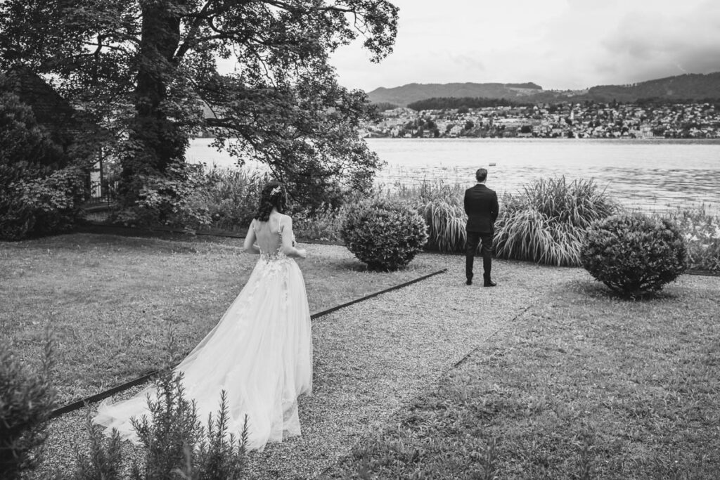 First Look in Schwarzweiss am Zürichsee Die Braut nähert sich dem Bräutigam von hinten beim First Look am Zürichsee – festgehalten in Schwarzweiss