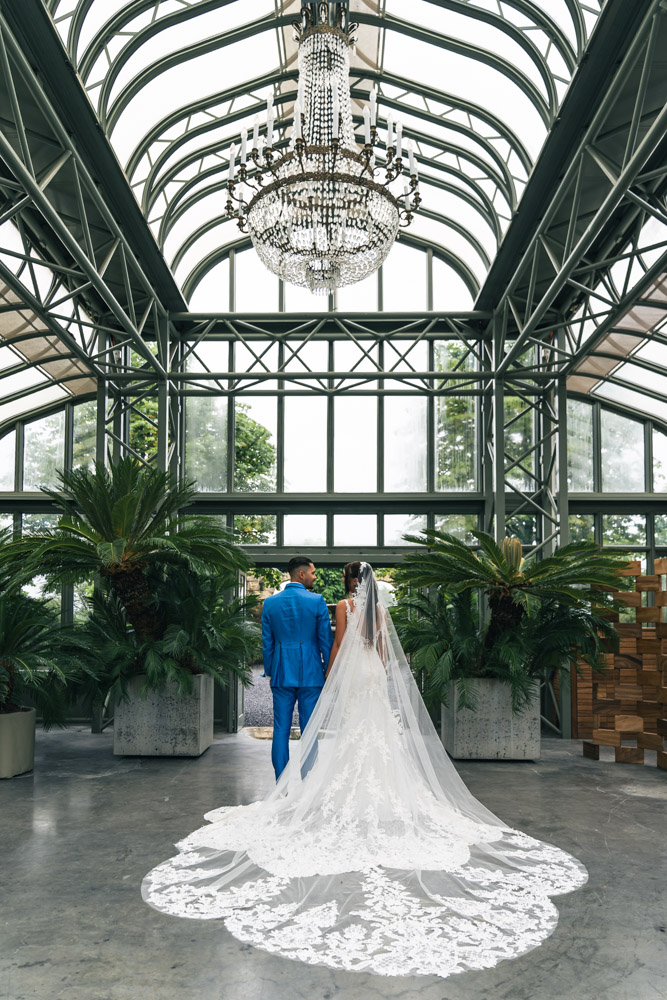 Hochzeit in der Enea Baumschule mit langem Schleier Brautpaar von hinten in der Enea Baumschule Rapperswil mit langem Schleier unter Glasdach und Kronleuchter – fotografiert von Christian Meixner.